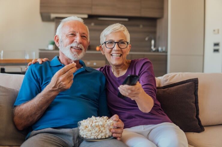 Coppia di anziani sorridenti con popcorn. Lei ha in mano il telecomando